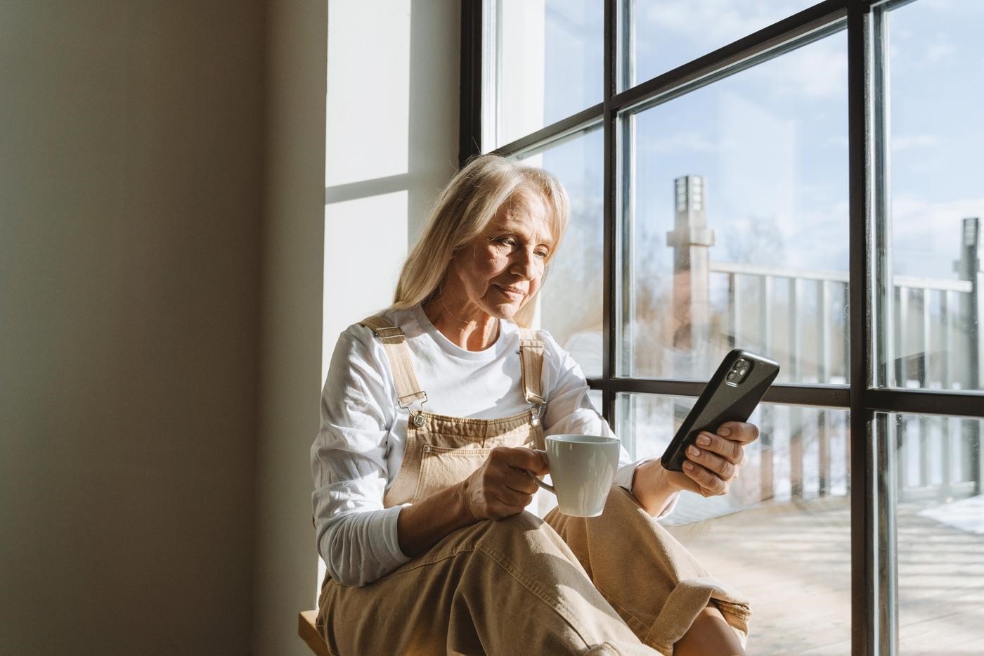 Older woman sitting next to a window checking her smartphone and drinking coffee